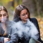 Vape lgbt teenagers. Bisexual lesbian young caucasian teenage girls in casual clothes vape electronic cigarette on the street in the park in the autumn evening. Bad habit.